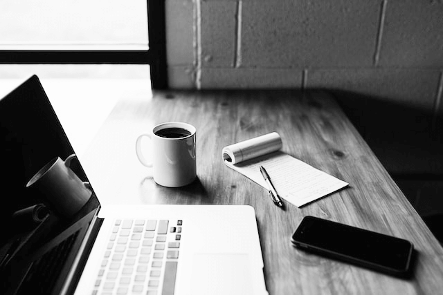 black and white image of a laptop, coffee cup, phone, and notepad